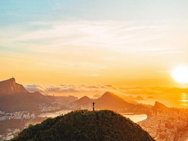 Person standing on a grassy hill with city and mountains at sunset.