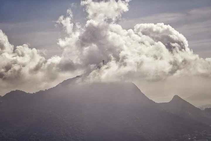 Christ the Redeemer statue barely visible atop mountain obscured by thick clouds.