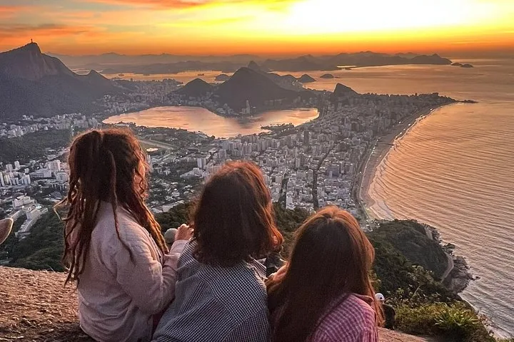 Three people sitting on a rock watching a colorful sunset over Rio de Janeiro.